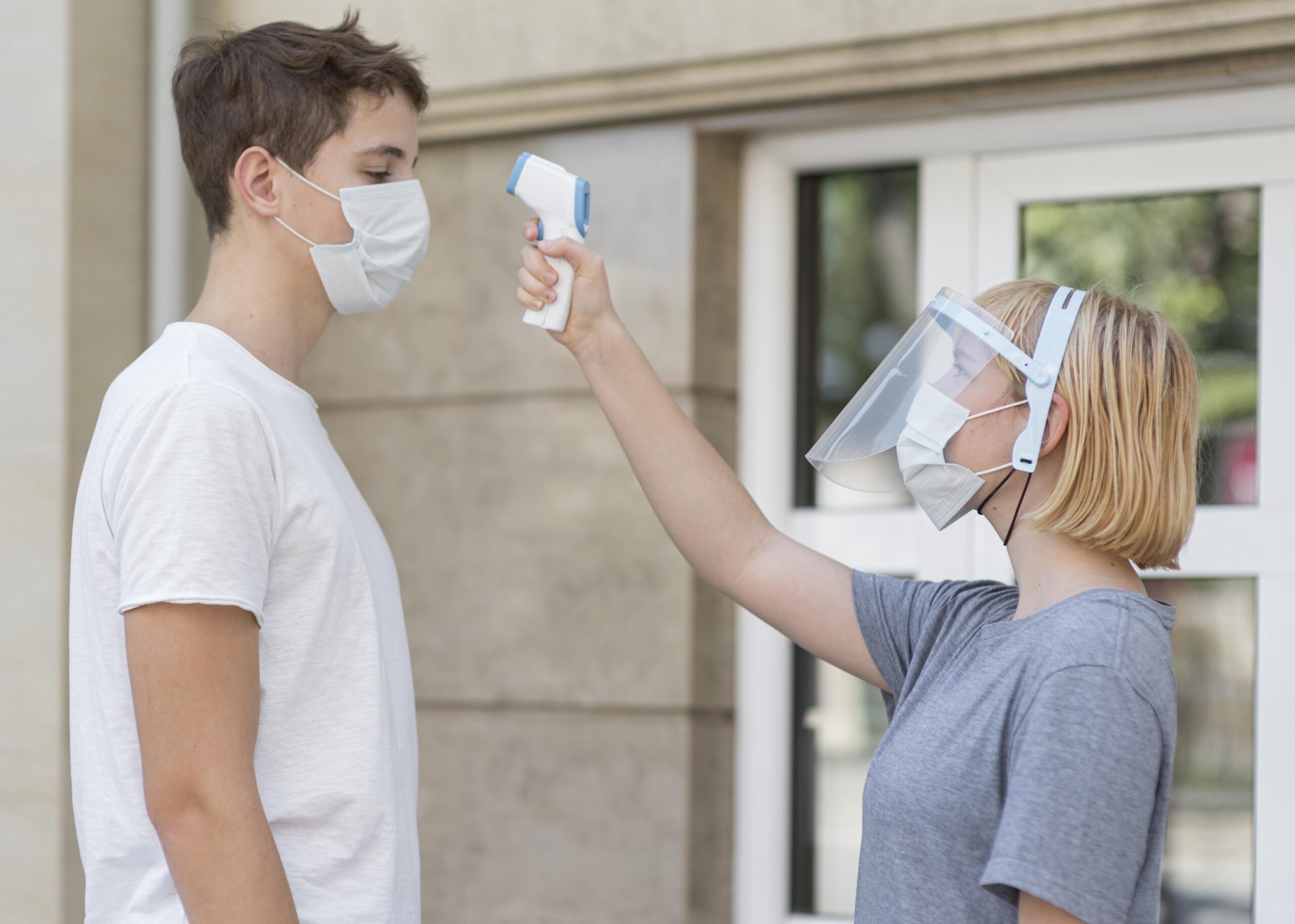 Person wearing a mask and face shield conducting a temperature check on another masked individual, emphasizing health and safety measures in mold inspection contexts.