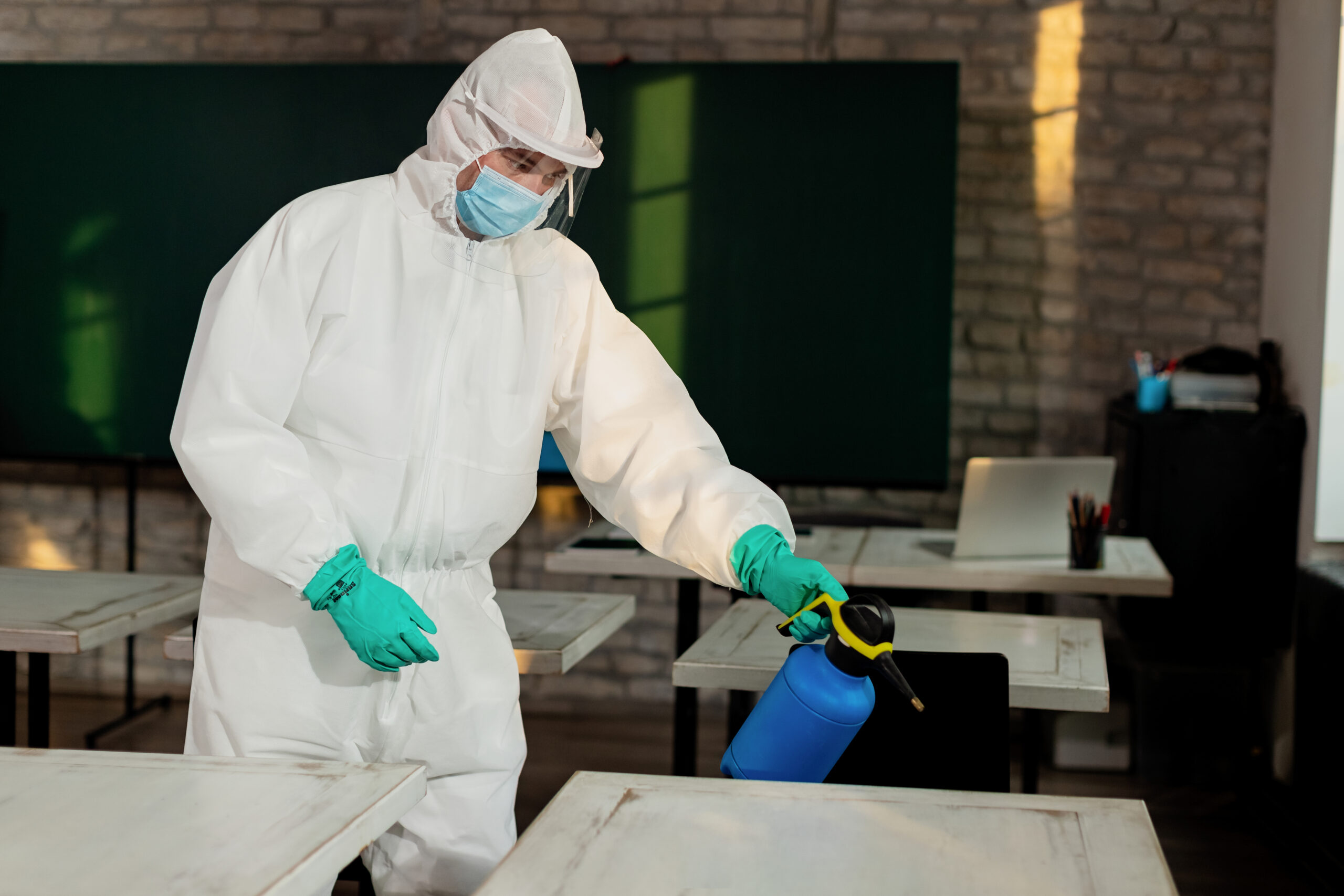 Man in hazmat suit disinfecting classroom surfaces with a spray bottle, emphasizing safety measures in mold prevention and indoor air quality management.