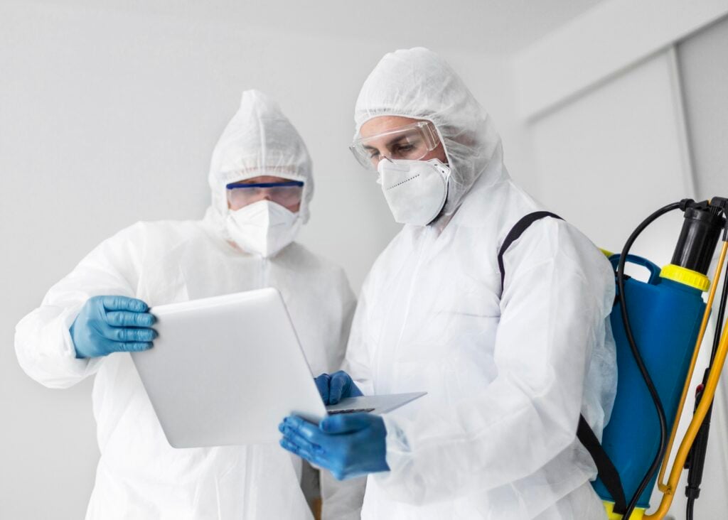 Two technicians in protective gear reviewing data on a laptop, equipped with mold remediation equipment, emphasizing professional mold inspection and remediation services.