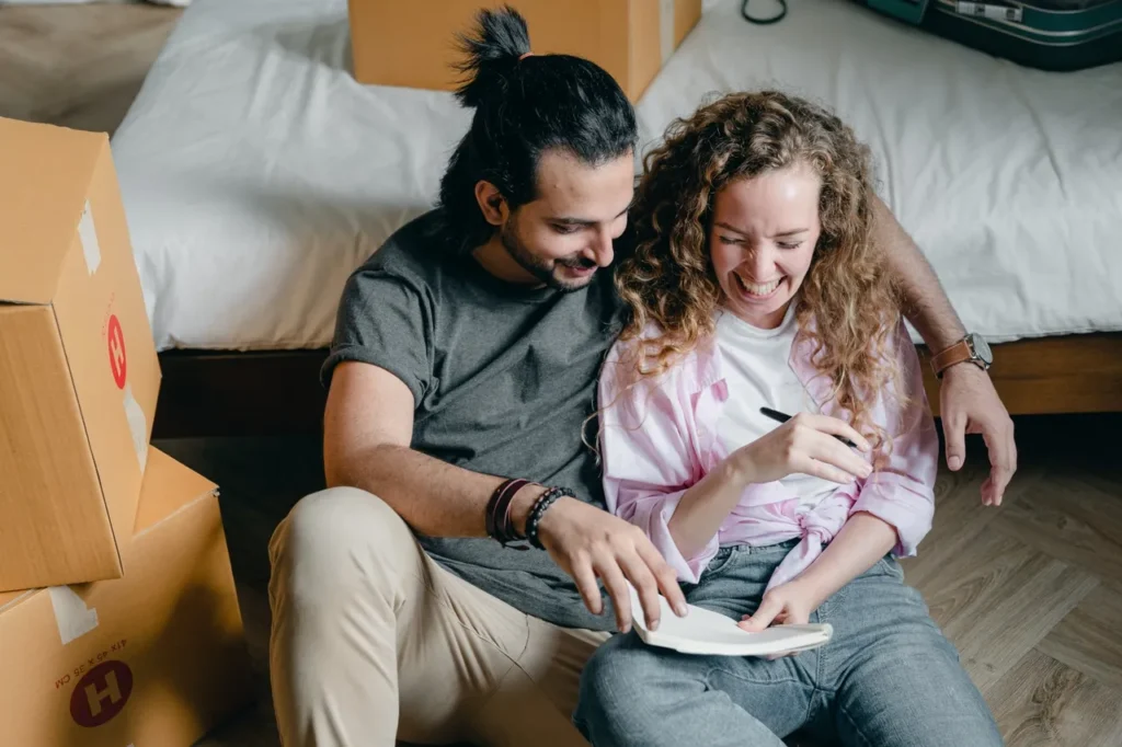 Couple sitting on the floor among moving boxes, smiling and engaging in conversation while reviewing notes, reflecting the positive experience of settling into a new home.